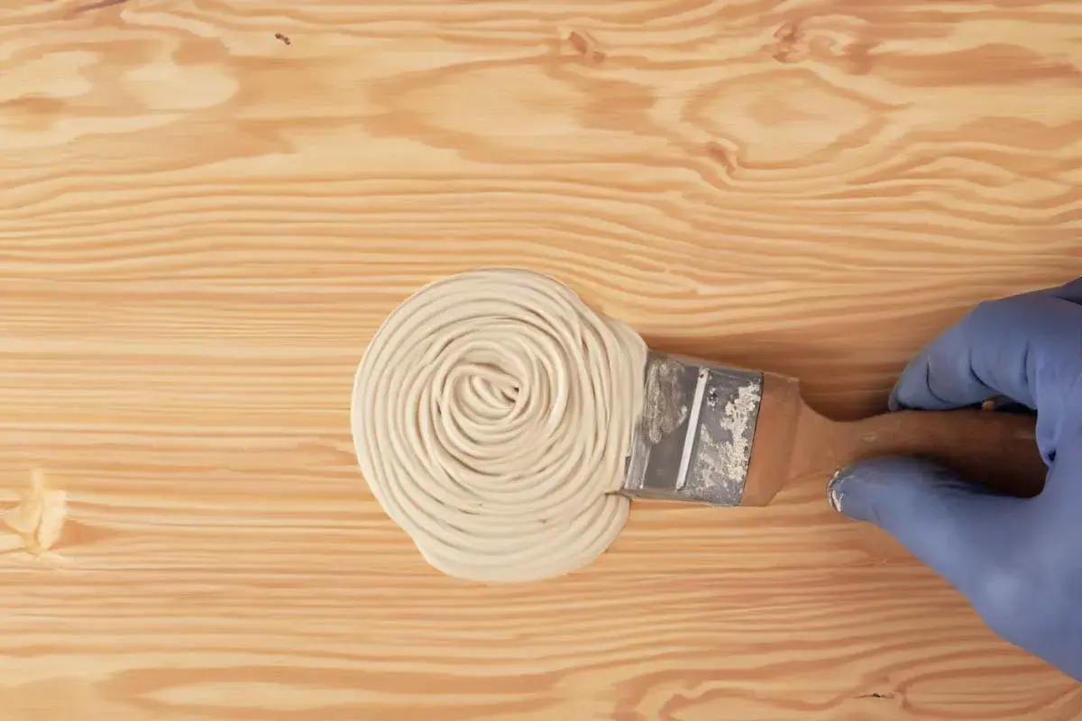 Mano con guanto blu applica stucco con pennello su legno. Guida pratica su come stuccare il legno per una finitura perfetta.