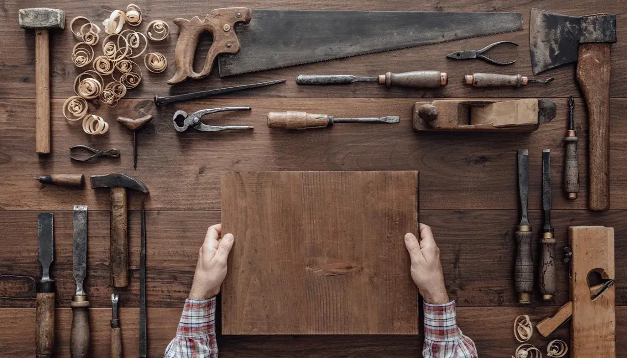 Mani che tengono un pezzo di legno, circondate da attrezzi da falegname, mostrando come si lavora il legno.