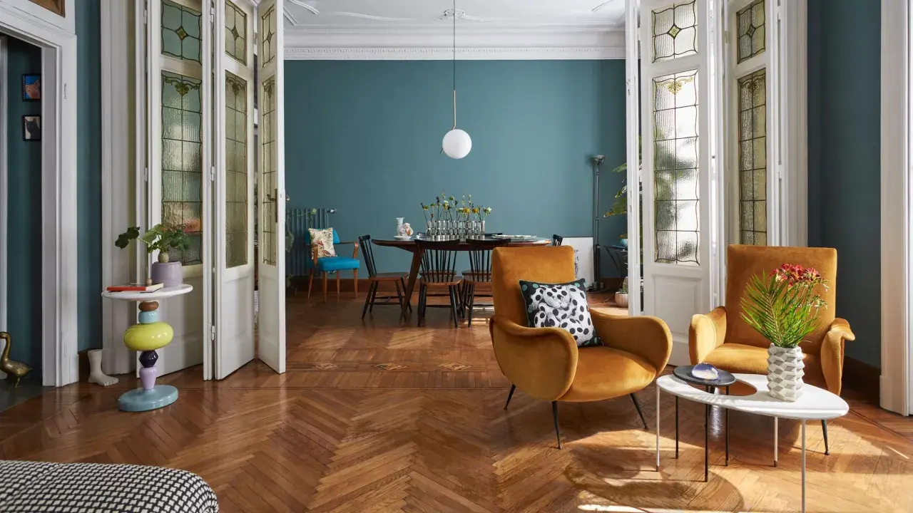 Soggiorno con poltrone ocra, tavolino moderno e un tavolo da pranzo in stile direttorio sullo sfondo. Porte a vetri decorati.