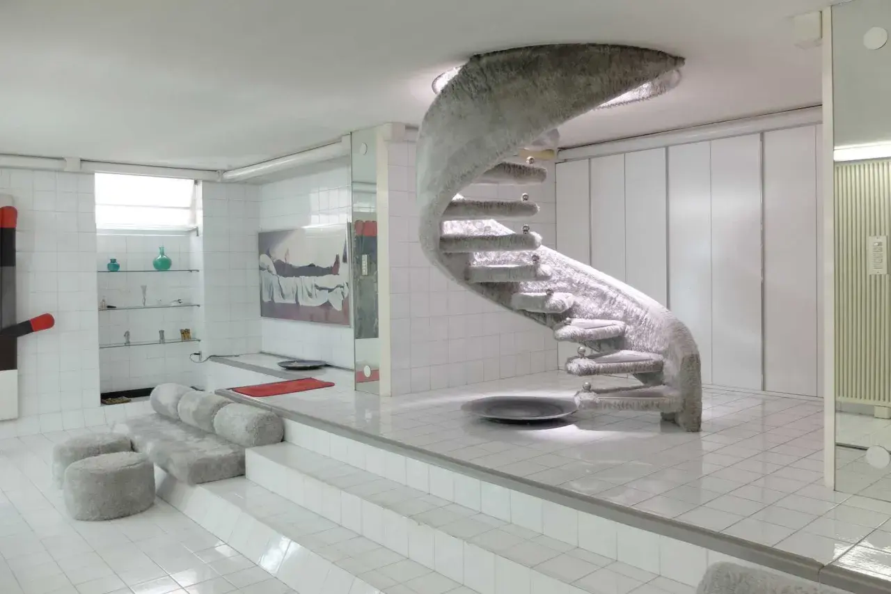 Interni futuristici con scala a chiocciola rivestita in pelliccia, un omaggio a Nanda Vigo. Divani e pouf morbidi completano l'ambiente.
