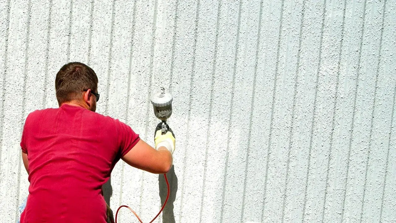 Uomo con maglietta rossa effettua verniciatura a spruzzo fai da te su una parete esterna bianca e rigata.