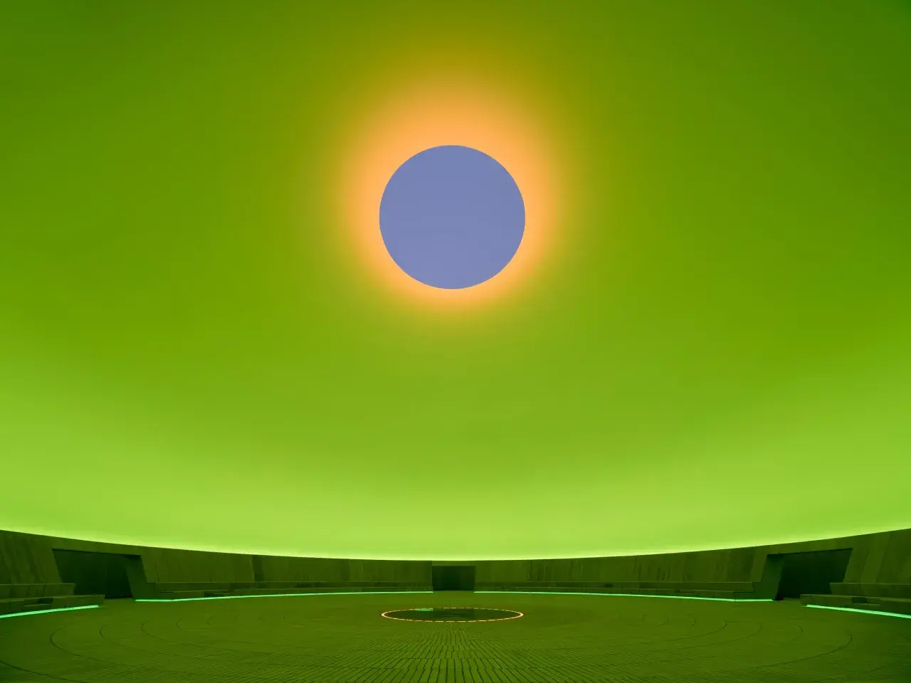 Interno di un'installazione di James Turrell: un cerchio blu su sfondo verde brillante, con un alone arancione. La sala circolare ha sedute e un cerchio luminoso al centro.