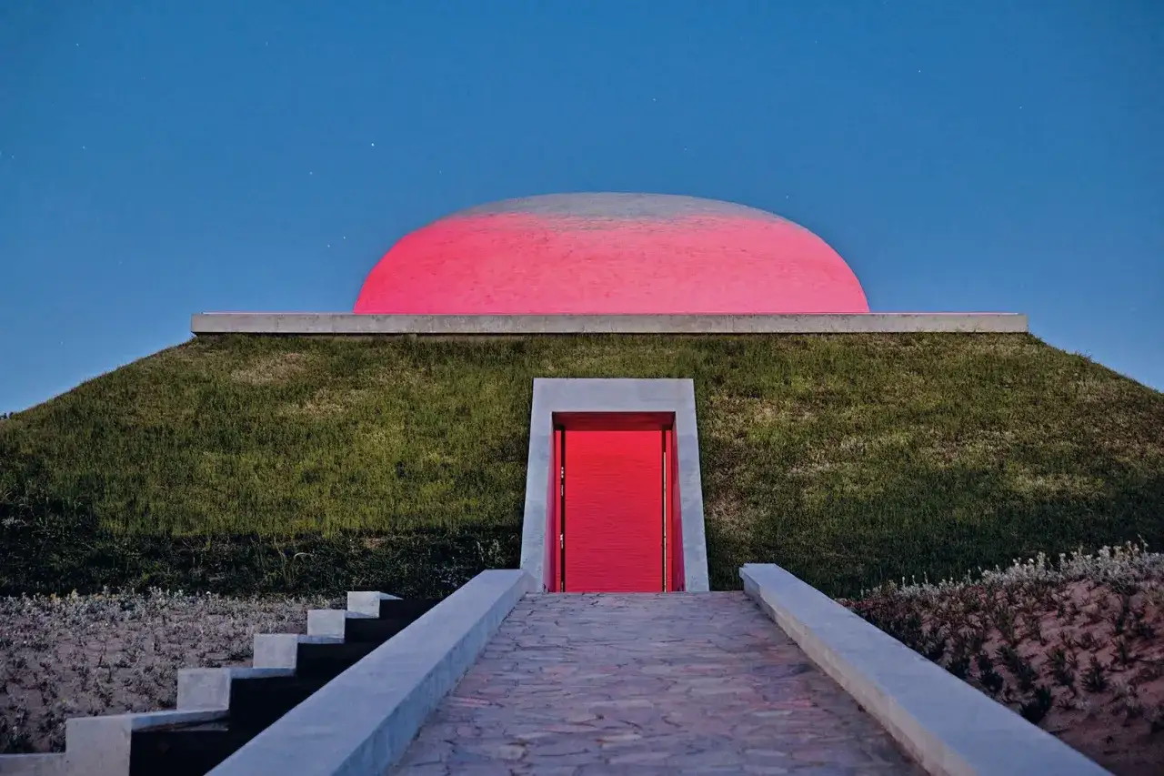 Cupola rosa brillante illumina un tumulo erboso, con una porta rossa che invita all'interno. Un'installazione di James Turrell.