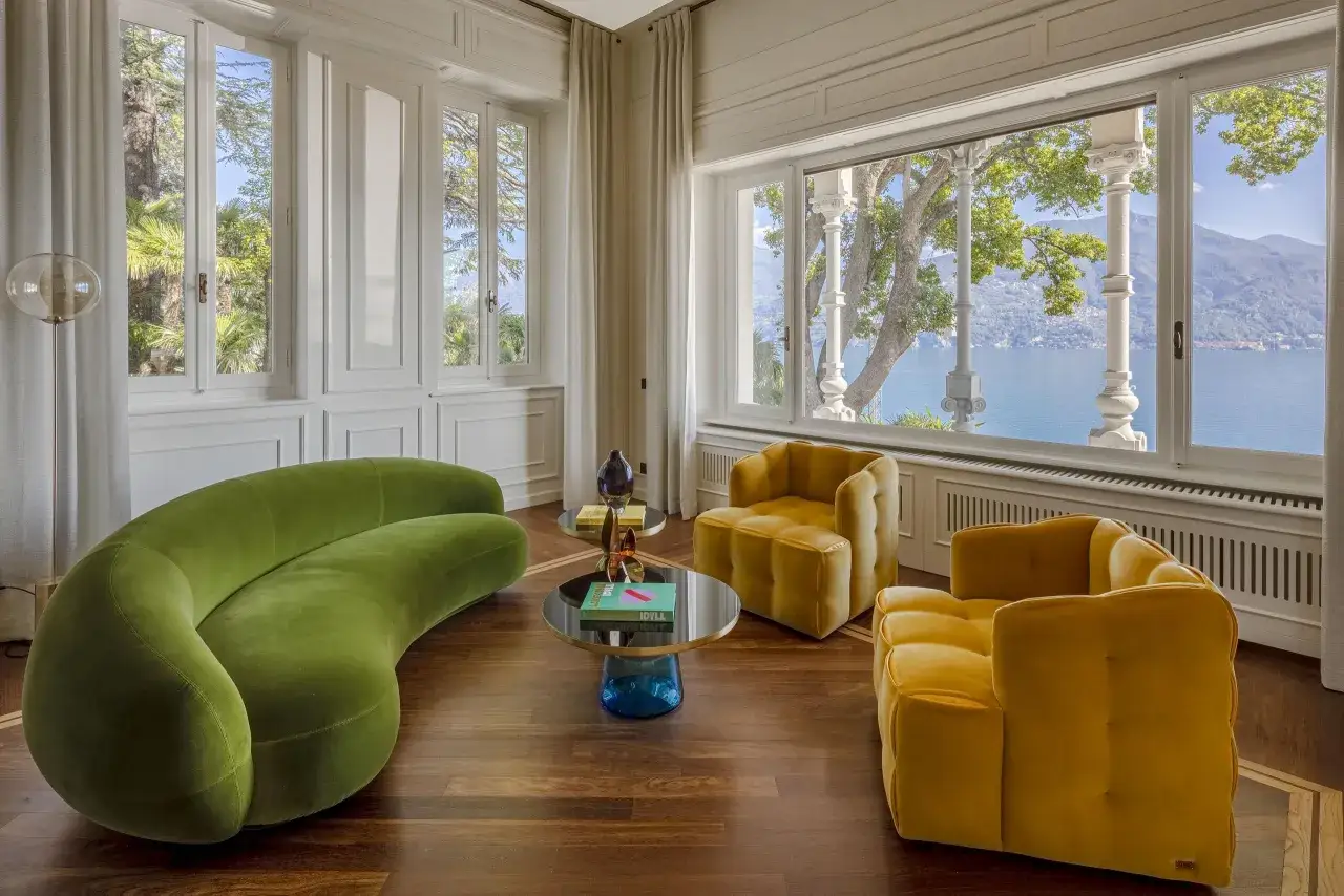 Divano verde sinuoso e poltrone gialle in velluto, con tavolini rotondi e vista lago. Stile moderno con richiami art deco.