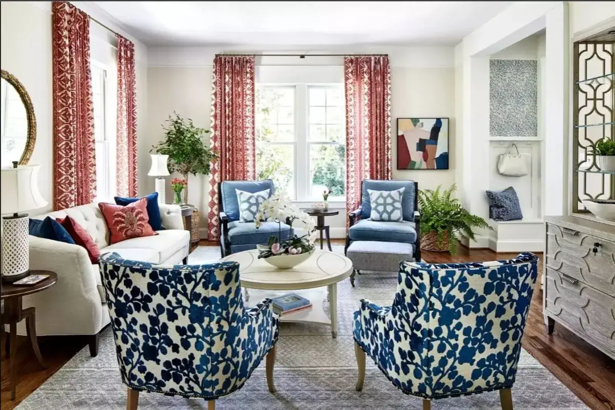 Salotto in stile grandmillennial con divano bianco, poltrone blu e sedie con fantasia floreale blu e bianca. Tende rosse con motivo geometrico.