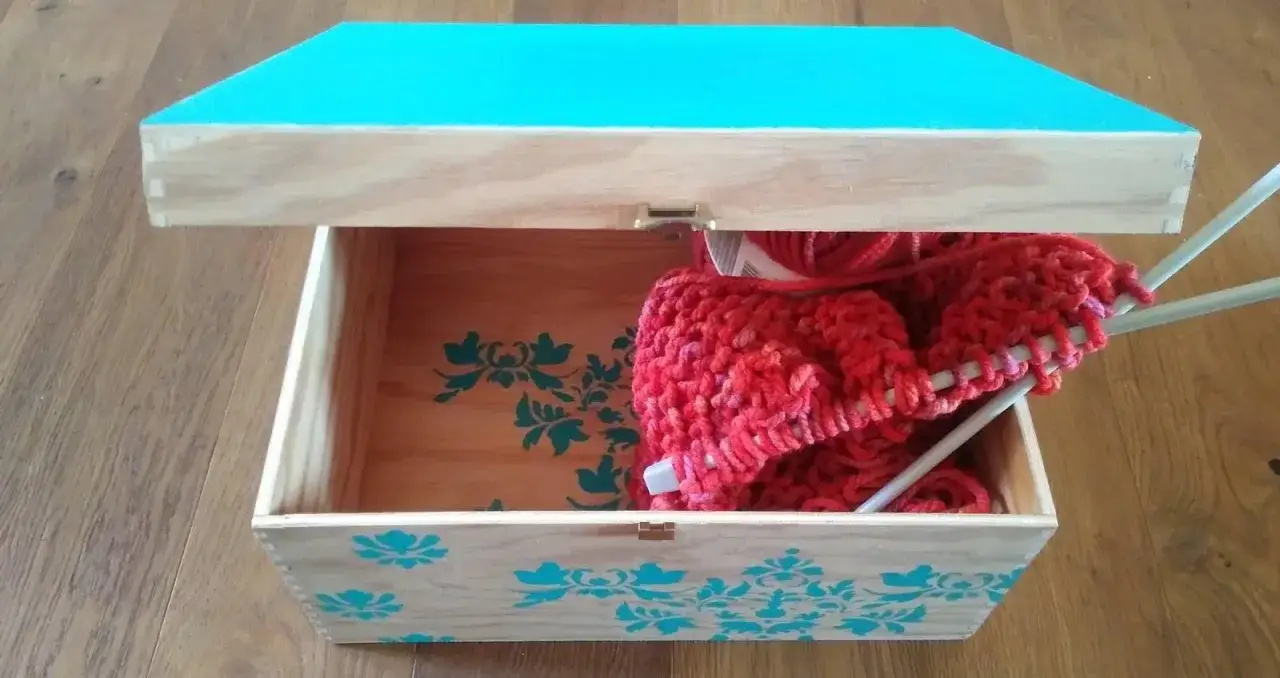 Scatole di legno decorate fai da te con motivi floreali turchesi, una contiene gomitoli rossi e ferri da maglia.