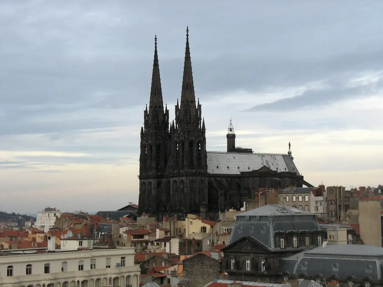 Imponente cattedrale gotica con torri aguzze che si stagliano su un cielo nuvoloso, dominando un paesaggio urbano di tetti rossi.