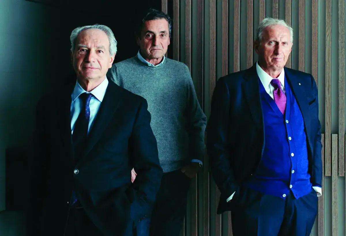Aldo Spinelli con due uomini eleganti, uno in maglione grigio, l'altro in abito blu e cardigan viola.