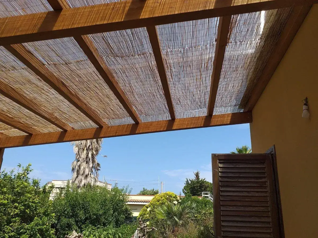 Tettoia fai da te in legno e canne, con vista su un giardino rigoglioso e cielo azzurro.