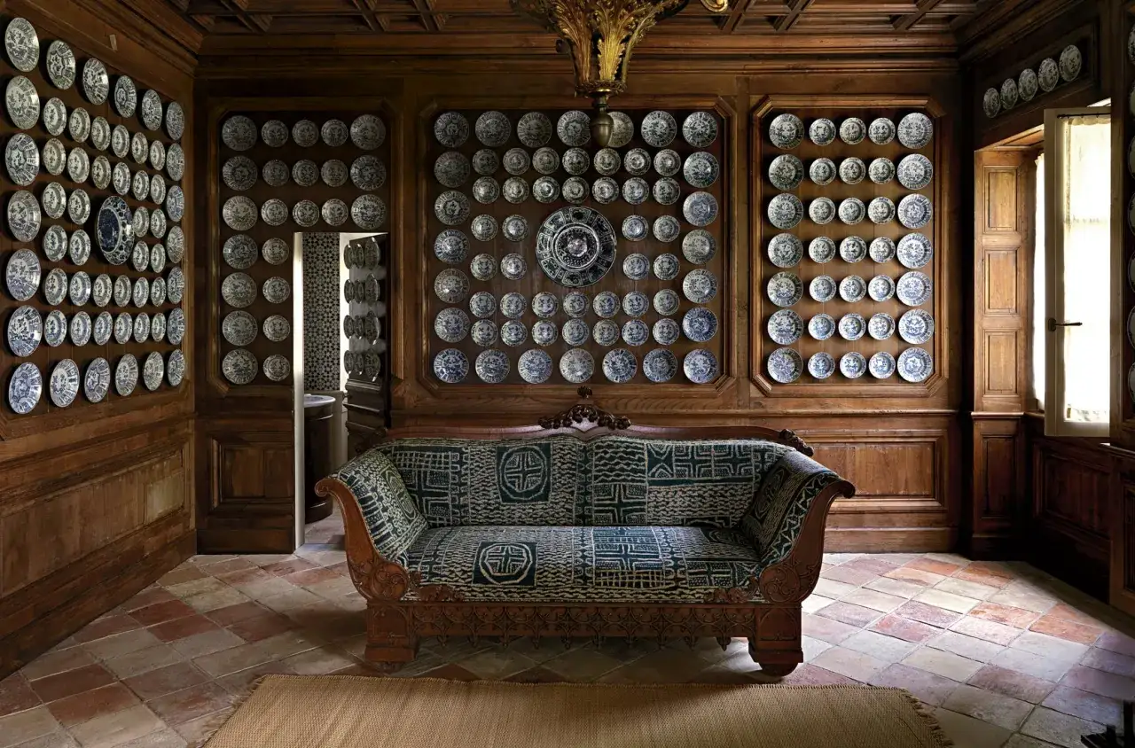 Interno con divano decorato e pareti rivestite di piatti in ceramica blu e bianchi, un'ambientazione che evoca lo stile dello studio Peregalli.