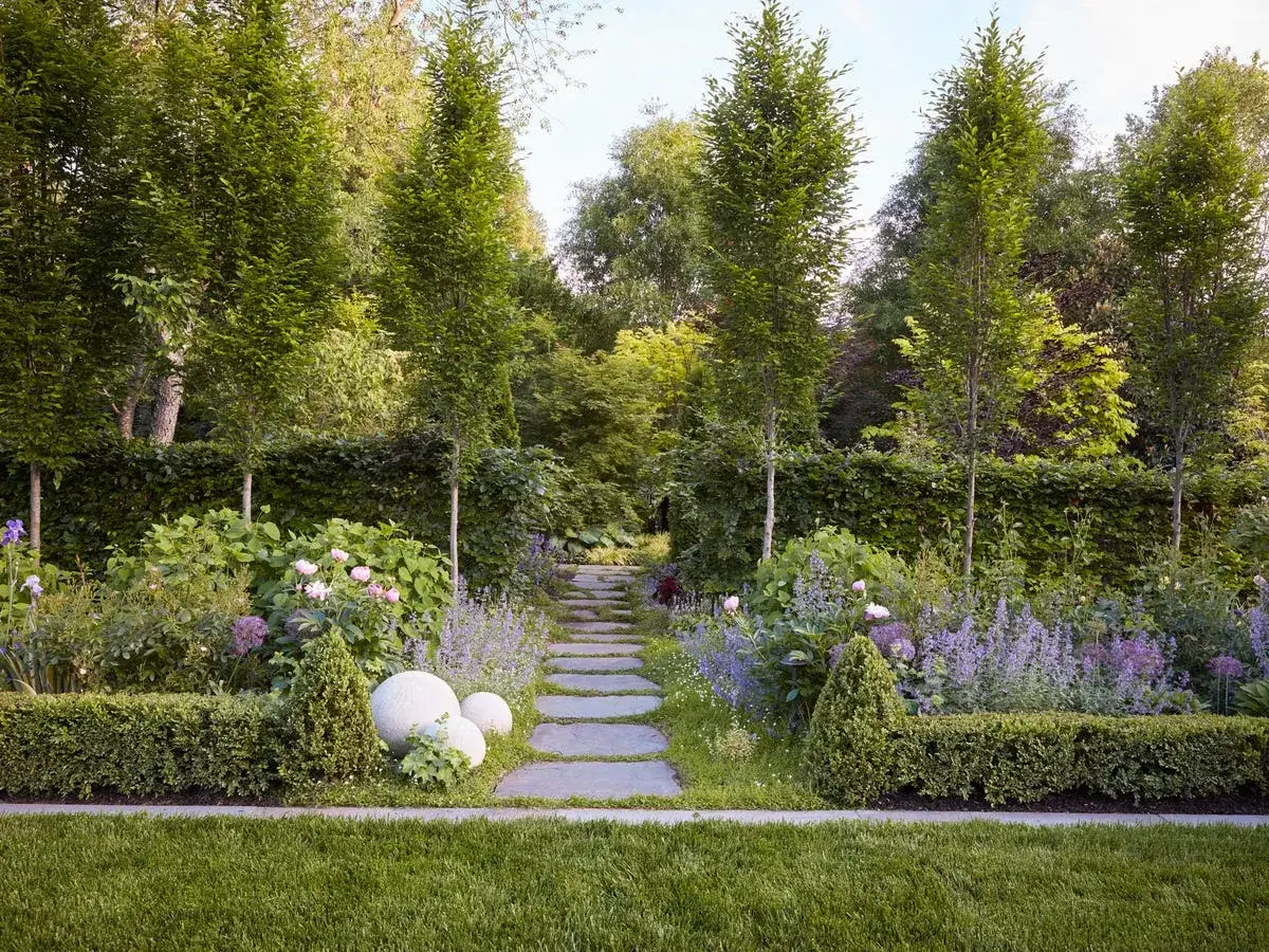 Percorso in pietra attraverso aiuole giardino idee con fiori viola e rosa, siepi curate e alberi slanciati.