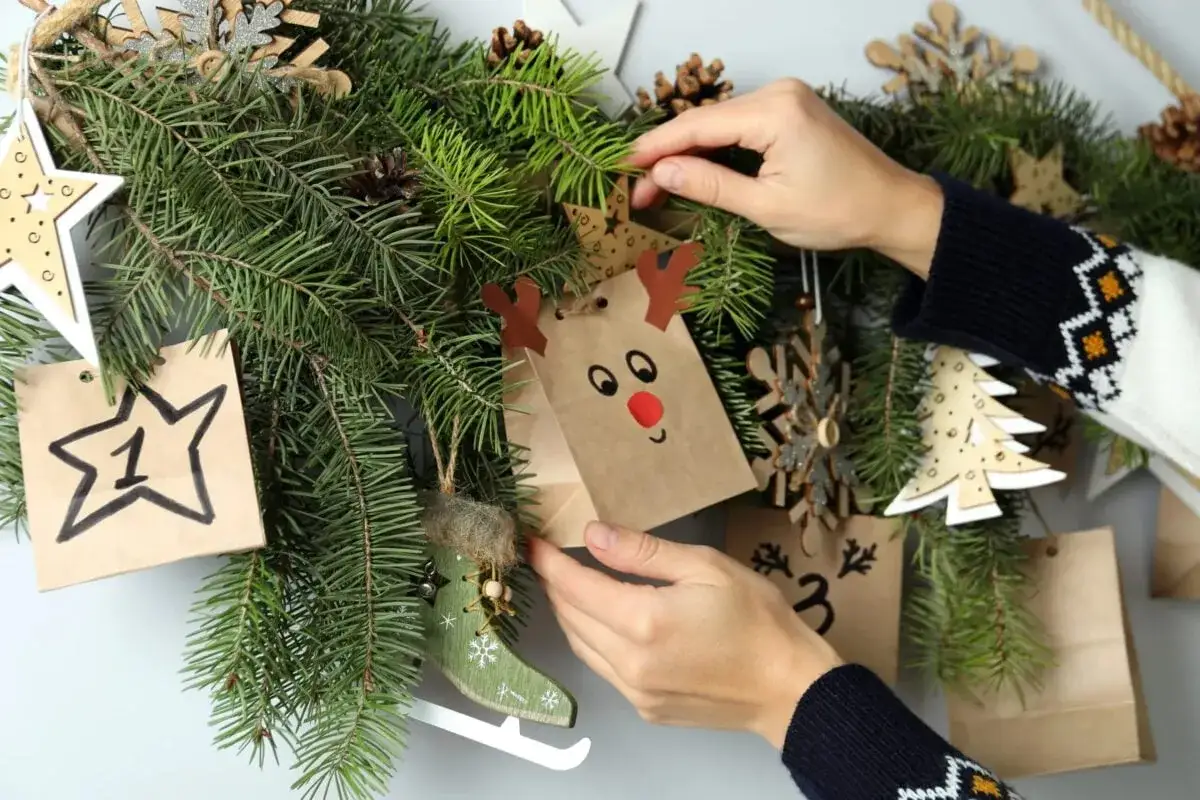 Mani che decorano un calendario dell'avvento con sacchetti natalizi fai da te, pigne e decorazioni in legno.